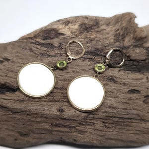May include: A pair of drop earrings featuring circular white discs set in gold-toned metal frames. Each earring is suspended from a gold-toned leverback closure by a small, round, light green gemstone accent positioned between the disc and the hook.