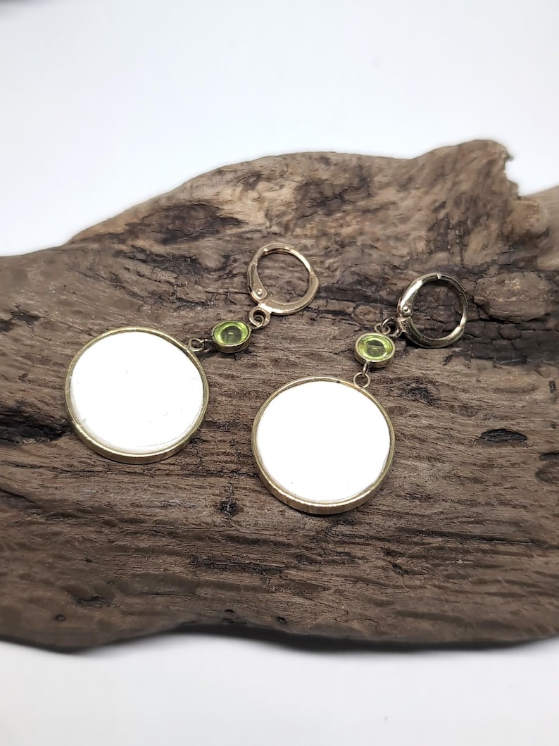 May include: A pair of drop earrings featuring circular white discs set in gold-toned metal frames. Each earring is suspended from a gold-toned leverback closure by a small, round, light green gemstone accent positioned between the disc and the hook.