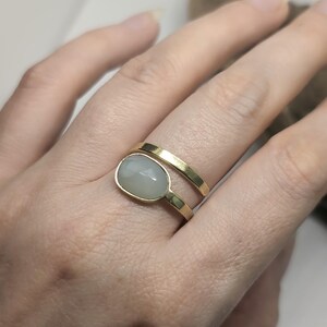 May include: A gold bypass ring featuring a smooth, polished gold band that wraps around the finger. One end of the band is plain, while the other end is set with a single, oval-shaped, translucent, pale grey cabochon gemstone in a gold bezel setting.