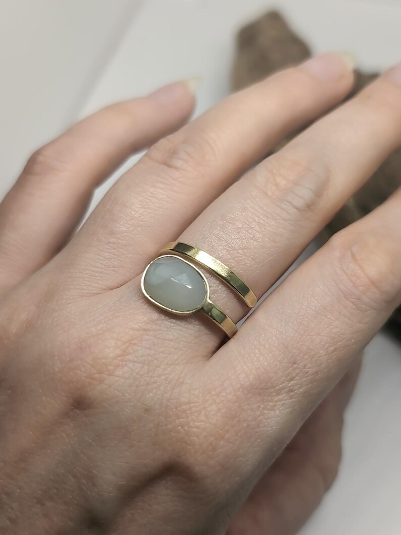 May include: A gold bypass ring featuring a smooth, polished gold band that wraps around the finger. One end of the band is plain, while the other end is set with a single, oval-shaped, translucent, pale grey cabochon gemstone in a gold bezel setting.