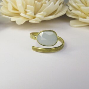 May include: An adjustable ring featuring a faceted, pale gray oval stone set in a brushed gold-tone metal band. The band has an open design that wraps around the finger, showcasing a matte finish on the metal surface and a smooth, polished stone.