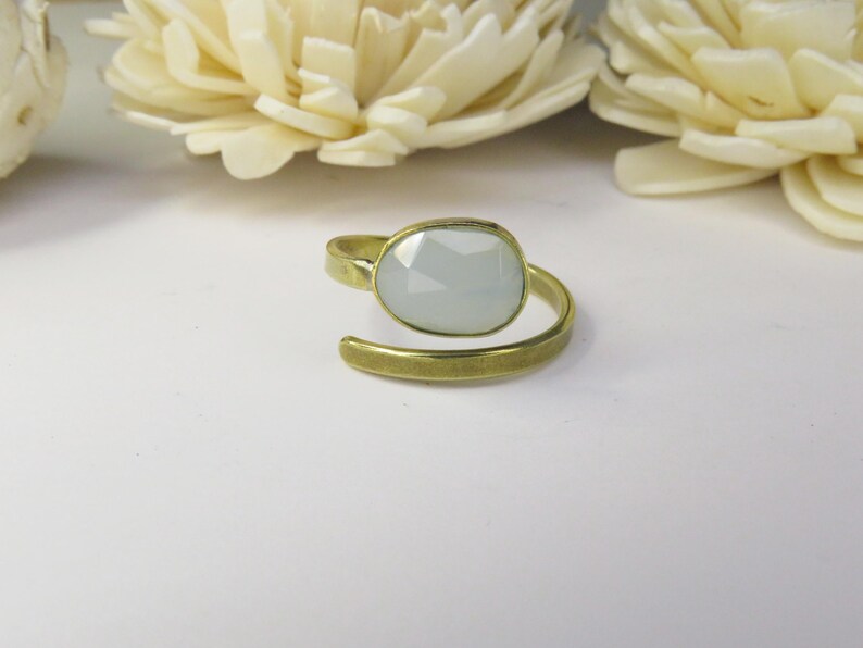 May include: An adjustable ring featuring a faceted, pale gray oval stone set in a brushed gold-tone metal band. The band has an open design that wraps around the finger, showcasing a matte finish on the metal surface and a smooth, polished stone.