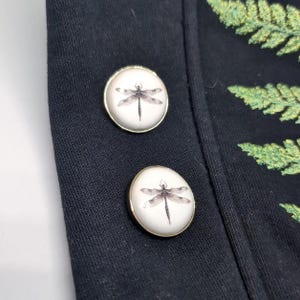 Op de afbeelding: Twee ronde knopen met een witte achtergrond en een zwarte libelle-illustratie, voorzien van een metalen goudkleurige rand. Deze knopen zijn bevestigd aan een zwart kledingstuk dat aan de rechterkant een gedeeltelijk groen varenbladpatroon vertoont.