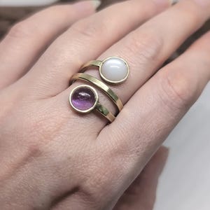 May include: A gold-toned bypass ring featuring two circular settings. One setting contains a smooth, opaque white cabochon, while the other setting holds a translucent, round purple gemstone. The band is open and wraps around the finger.