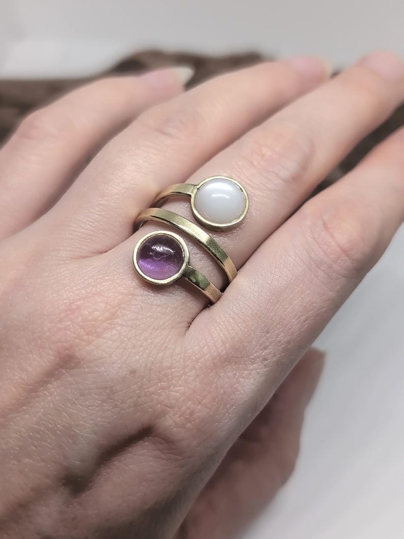 May include: A gold-toned bypass ring featuring two circular settings. One setting contains a smooth, opaque white cabochon, while the other setting holds a translucent, round purple gemstone. The band is open and wraps around the finger.