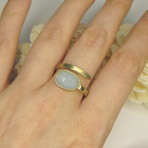 May include: A gold-toned bypass ring featuring a smooth, oval-shaped, pale blue cabochon stone set in a bezel. The ring band has a matte finish and wraps around the finger with two open ends, showcasing a clean, simple design with a soft color palette.