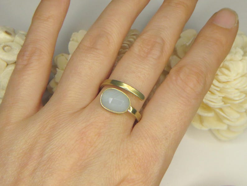 May include: A gold-toned bypass ring featuring a smooth, oval-shaped, pale blue cabochon stone set in a bezel. The ring band has a matte finish and wraps around the finger with two open ends, showcasing a clean, simple design with a soft color palette.