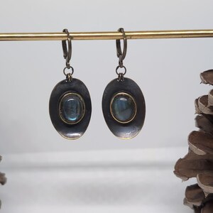 May include: A pair of drop earrings featuring oval-shaped, dark metallic settings with a distressed finish. Each earring contains a central, smooth, oval-cut labradorite gemstone displaying blue iridescence, suspended from a hinged lever-back clasp.