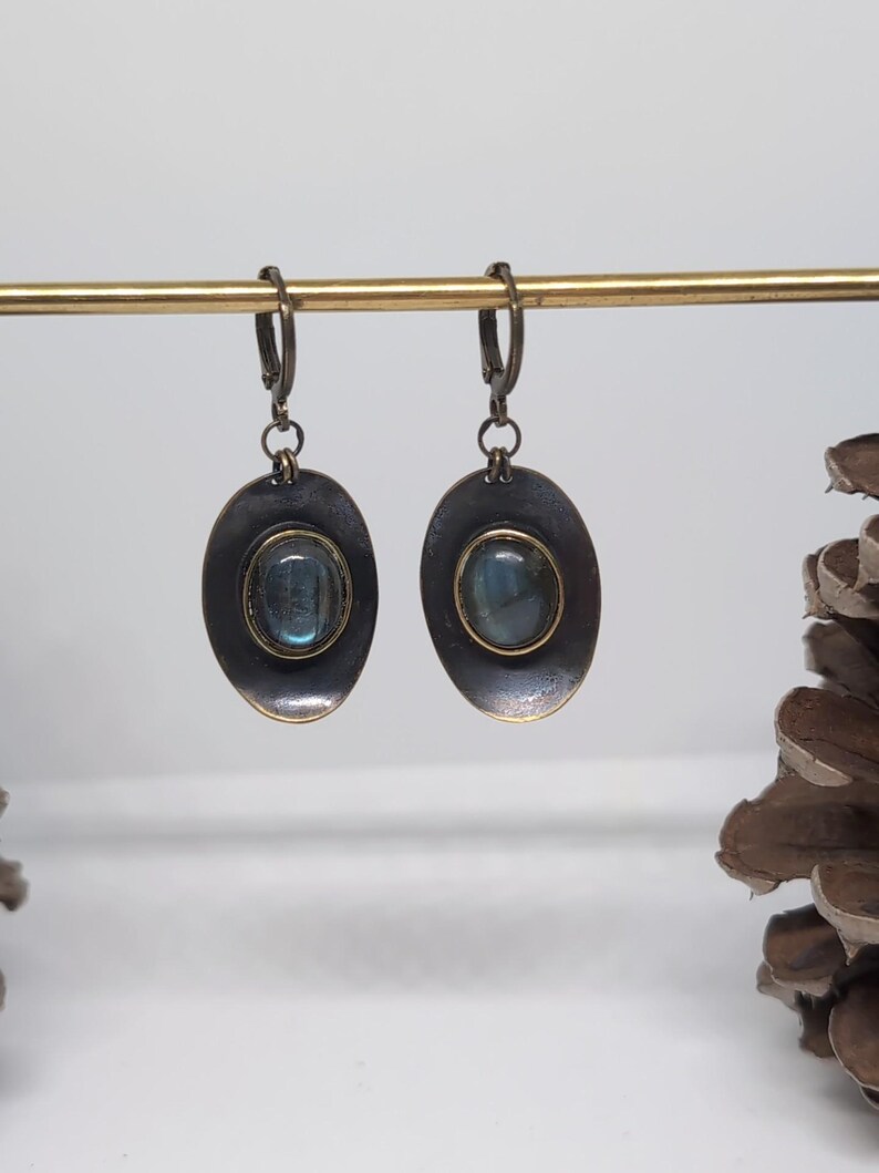 May include: A pair of drop earrings featuring oval-shaped, dark metallic settings with a distressed finish. Each earring contains a central, smooth, oval-cut labradorite gemstone displaying blue iridescence, suspended from a hinged lever-back clasp.