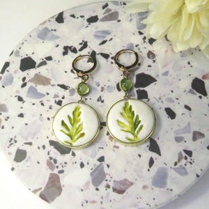 May include: A pair of gold-tone hoop earrings featuring a small green circular bead and a larger white circular pendant with a painted green leaf design. The earrings have a hinged closure and are displayed as a matching set of two identical pieces.