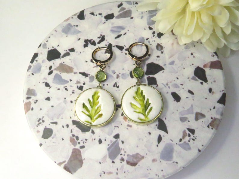 May include: A pair of gold-tone hoop earrings featuring a small green circular bead and a larger white circular pendant with a painted green leaf design. The earrings have a hinged closure and are displayed as a matching set of two identical pieces.