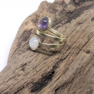 May include: A bypass ring featuring a gold-toned metal band that spirals around the finger. The ring is set with two round cabochon stones: one is a translucent purple amethyst and the other is an opaque white moonstone or similar gem.