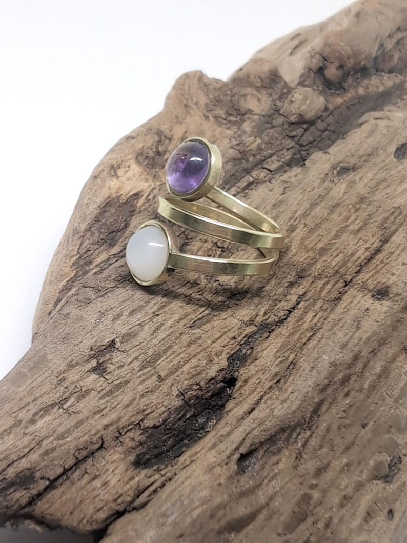 May include: A bypass ring featuring a gold-toned metal band that spirals around the finger. The ring is set with two round cabochon stones: one is a translucent purple amethyst and the other is an opaque white moonstone or similar gem.