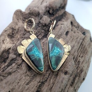 May include: A pair of dangle earrings featuring triangular-shaped stones with dark green and teal marbled patterns. The stones are set in textured gold-toned metal frames with a fan-like edge and attached to small circular hinged hoop closures.