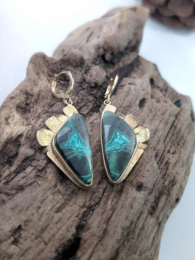 May include: A pair of dangle earrings featuring triangular-shaped stones with dark green and teal marbled patterns. The stones are set in textured gold-toned metal frames with a fan-like edge and attached to small circular hinged hoop closures.