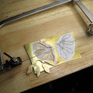 May include: A close-up of a metal cutting project. A piece of brass is being cut with a scroll saw. The brass has two designs cut into it, one of a leaf and one of a flower. The scroll saw is attached to a wooden table.