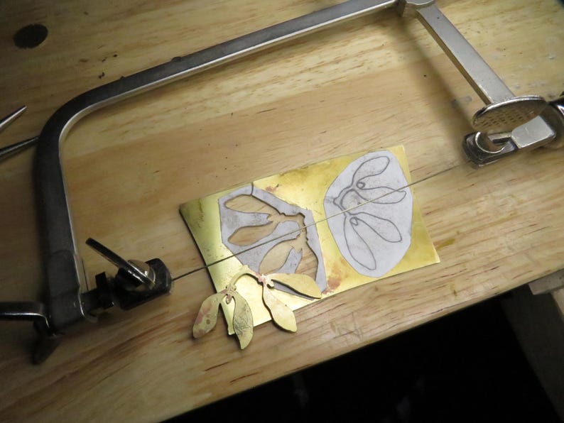 May include: A close-up of a metal cutting project. A piece of brass is being cut with a scroll saw. The brass has two designs cut into it, one of a leaf and one of a flower. The scroll saw is attached to a wooden table.
