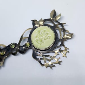 May include: A metal hair accessory featuring a dark, oxidized finish with an intricate, branch-like structure. It is adorned with small, light green leaves, two small round green gemstones, and a central, circular pale yellow cabochon.