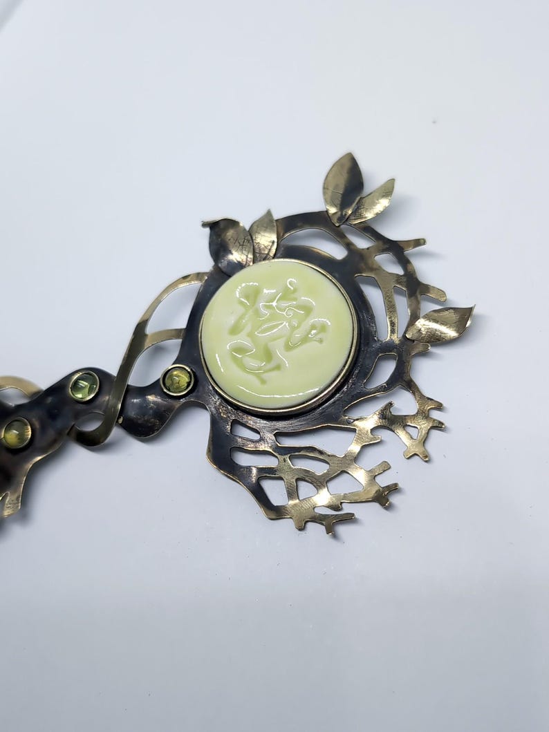 May include: A metal hair accessory featuring a dark, oxidized finish with an intricate, branch-like structure. It is adorned with small, light green leaves, two small round green gemstones, and a central, circular pale yellow cabochon.