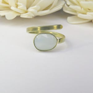 May include: A bypass ring featuring a gold-toned metal band with a matte finish and a single oval-shaped, faceted, translucent white stone set at the center. The band wraps around the finger in an open, adjustable design with smooth edges.