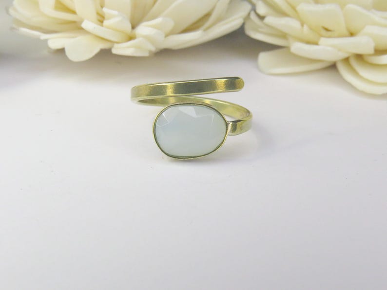 May include: A bypass ring featuring a gold-toned metal band with a matte finish and a single oval-shaped, faceted, translucent white stone set at the center. The band wraps around the finger in an open, adjustable design with smooth edges.