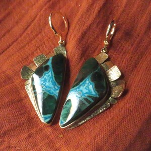 May include: A pair of dangle earrings featuring polished, triangular-shaped stones with deep green and vibrant turquoise patterns. Each stone is set in a textured, metallic frame with a fan-like arrangement of small, hammered gold-tone metal plates.