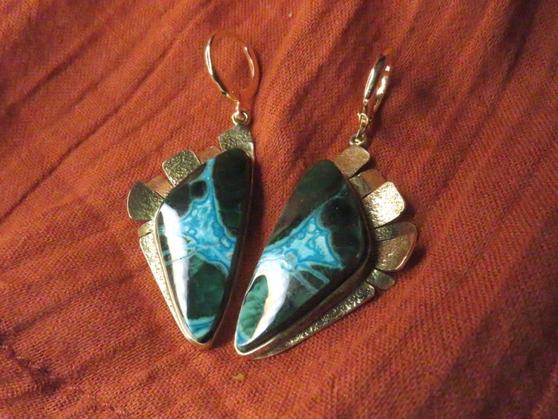 May include: A pair of dangle earrings featuring polished, triangular-shaped stones with deep green and vibrant turquoise patterns. Each stone is set in a textured, metallic frame with a fan-like arrangement of small, hammered gold-tone metal plates.