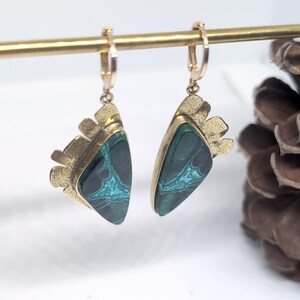 May include: A pair of dangle earrings featuring triangular dark green stones with intricate teal veining. Each stone is set in a textured gold-tone metal frame with a fan-like top edge, suspended from a small gold-tone hinged hoop earring component.