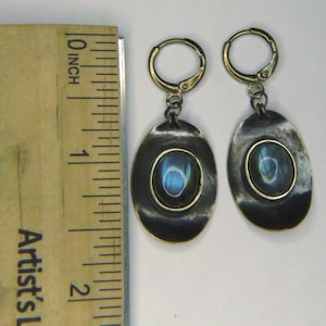 May include: A pair of oval-shaped drop earrings featuring dark, oxidized metal settings with a central iridescent blue stone. The earrings are positioned next to a wooden ruler, showing each earring measures approximately 1.5 inches in length.