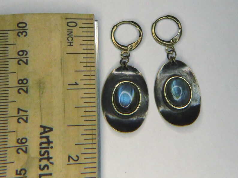 May include: A pair of oval-shaped drop earrings featuring dark, oxidized metal settings with a central iridescent blue stone. The earrings are positioned next to a wooden ruler, showing each earring measures approximately 1.5 inches in length.