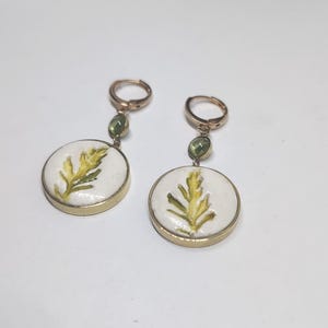 May include: A pair of gold-toned hoop earrings featuring a small green gemstone accent and a circular white pendant. Each pendant displays a painted yellow and green fern-like leaf design encased in a thin gold-colored metal rim.