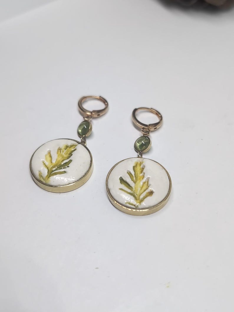 May include: A pair of gold-toned hoop earrings featuring a small green gemstone accent and a circular white pendant. Each pendant displays a painted yellow and green fern-like leaf design encased in a thin gold-colored metal rim.
