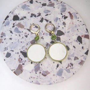 May include: A pair of gold-tone hoop earrings featuring a small green circular accent stone above a larger, flat, white circular pendant. The earrings have a hinged clasp mechanism and are constructed with a metallic gold-colored frame.