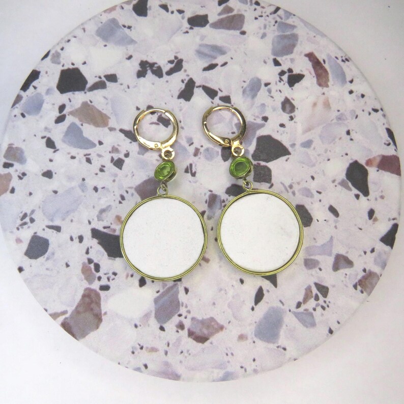 May include: A pair of gold-tone hoop earrings featuring a small green circular accent stone above a larger, flat, white circular pendant. The earrings have a hinged clasp mechanism and are constructed with a metallic gold-colored frame.