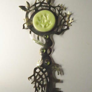 May include: A metal pendant necklace featuring a tree-like structure with a circular pale yellow center piece. The design includes intricate branch-like cutouts, three small green circular gemstones set vertically, and a thin metal chain.