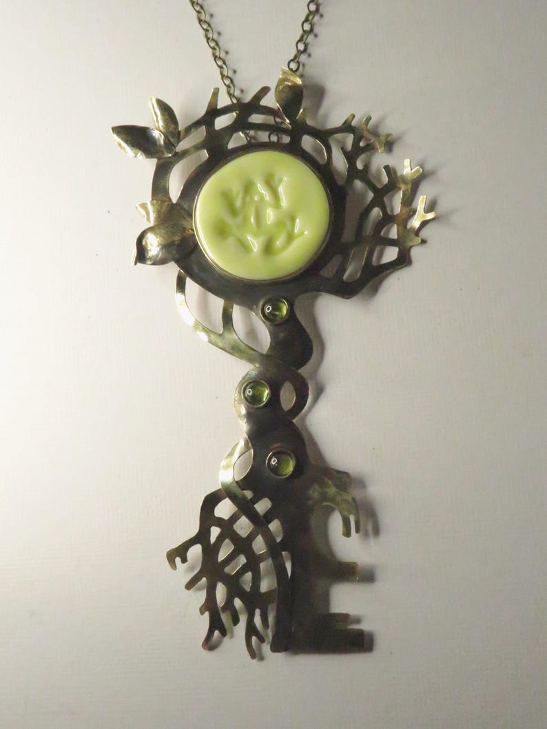 May include: A metal pendant necklace featuring a tree-like structure with a circular pale yellow center piece. The design includes intricate branch-like cutouts, three small green circular gemstones set vertically, and a thin metal chain.