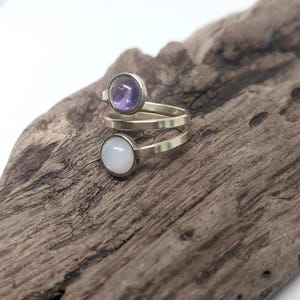 May include: A bypass ring featuring a metallic band that spirals around the finger. The ring is adorned with two circular cabochon stones: one is a translucent purple amethyst, and the other is an opaque, smooth white moonstone or opal-like gem.