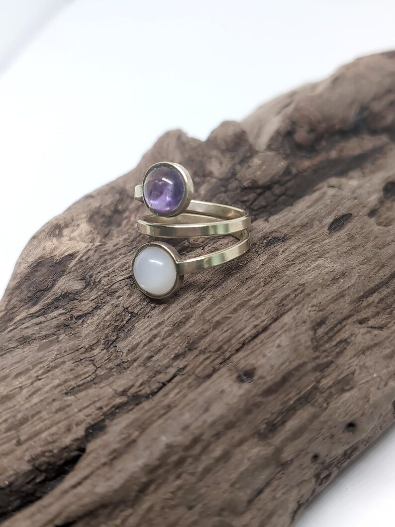 May include: A bypass ring featuring a metallic band that spirals around the finger. The ring is adorned with two circular cabochon stones: one is a translucent purple amethyst, and the other is an opaque, smooth white moonstone or opal-like gem.