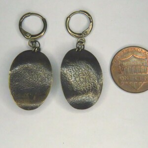 May include: A pair of bronze-toned oval drop earrings with a textured, dark patina finish and hinged hoop closures, displayed next to a United States one-cent coin for scale to show the size of the jewelry pieces.