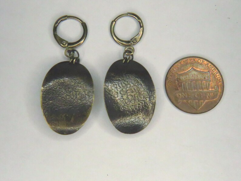 May include: A pair of bronze-toned oval drop earrings with a textured, dark patina finish and hinged hoop closures, displayed next to a United States one-cent coin for scale to show the size of the jewelry pieces.