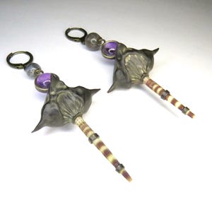 May include: A pair of unique earrings featuring a dark gray, stylized flower design. The earrings have a purple bead and a striped, cylindrical stem. The earrings are attached to a bronze-colored hoop.