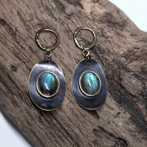 May include: A pair of drop earrings featuring oval-shaped labradorite gemstones with blue and green iridescence. Each stone is set in a dark, oxidized metal frame with a smooth, rounded edge, suspended from a brass-toned hinged hoop closure.