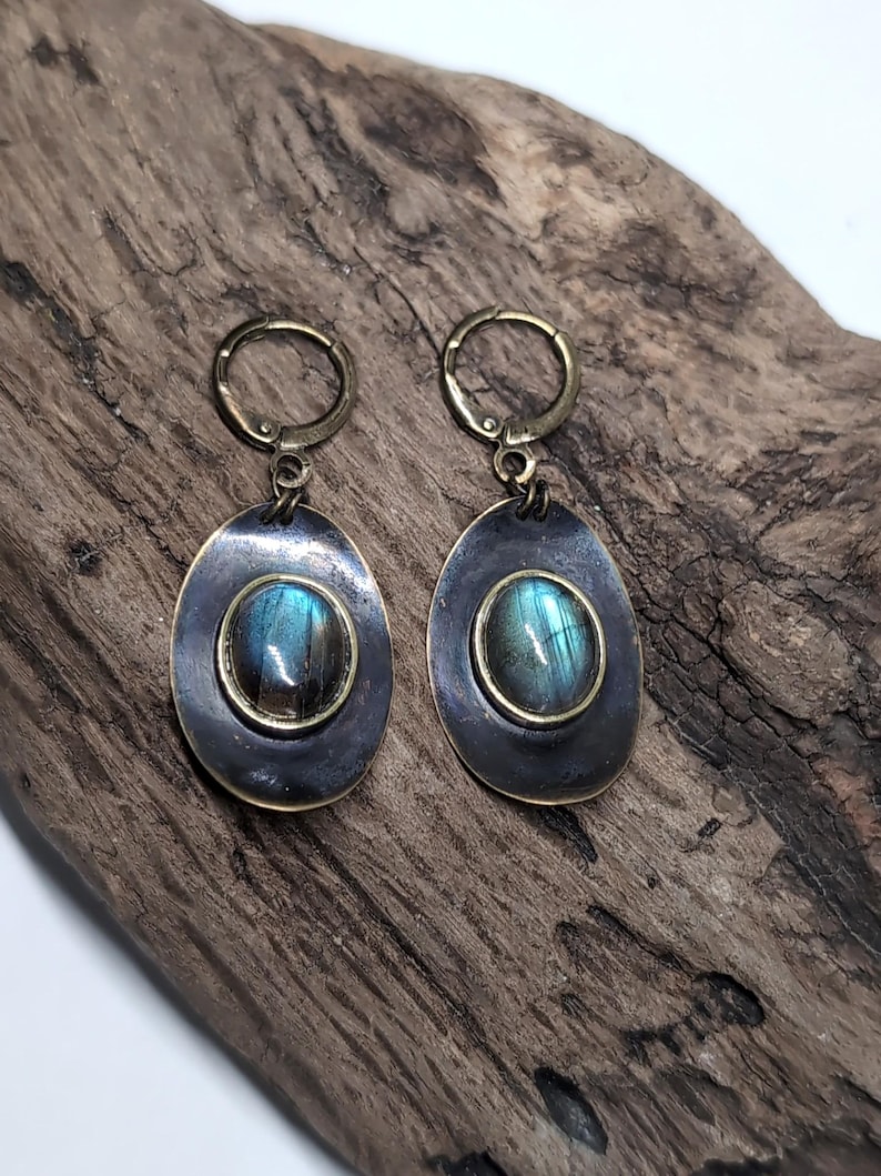 May include: A pair of drop earrings featuring oval-shaped labradorite gemstones with blue and green iridescence. Each stone is set in a dark, oxidized metal frame with a smooth, rounded edge, suspended from a brass-toned hinged hoop closure.