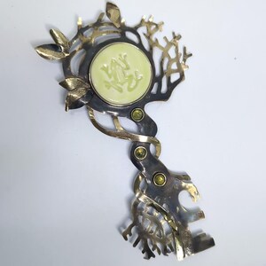 May include: A decorative metal key-shaped brooch featuring a central pale yellow circular inlay with a relief of a frog. The metalwork has a dark, oxidized finish with polished gold-tone leaf accents and three small, round, green inset gemstones.