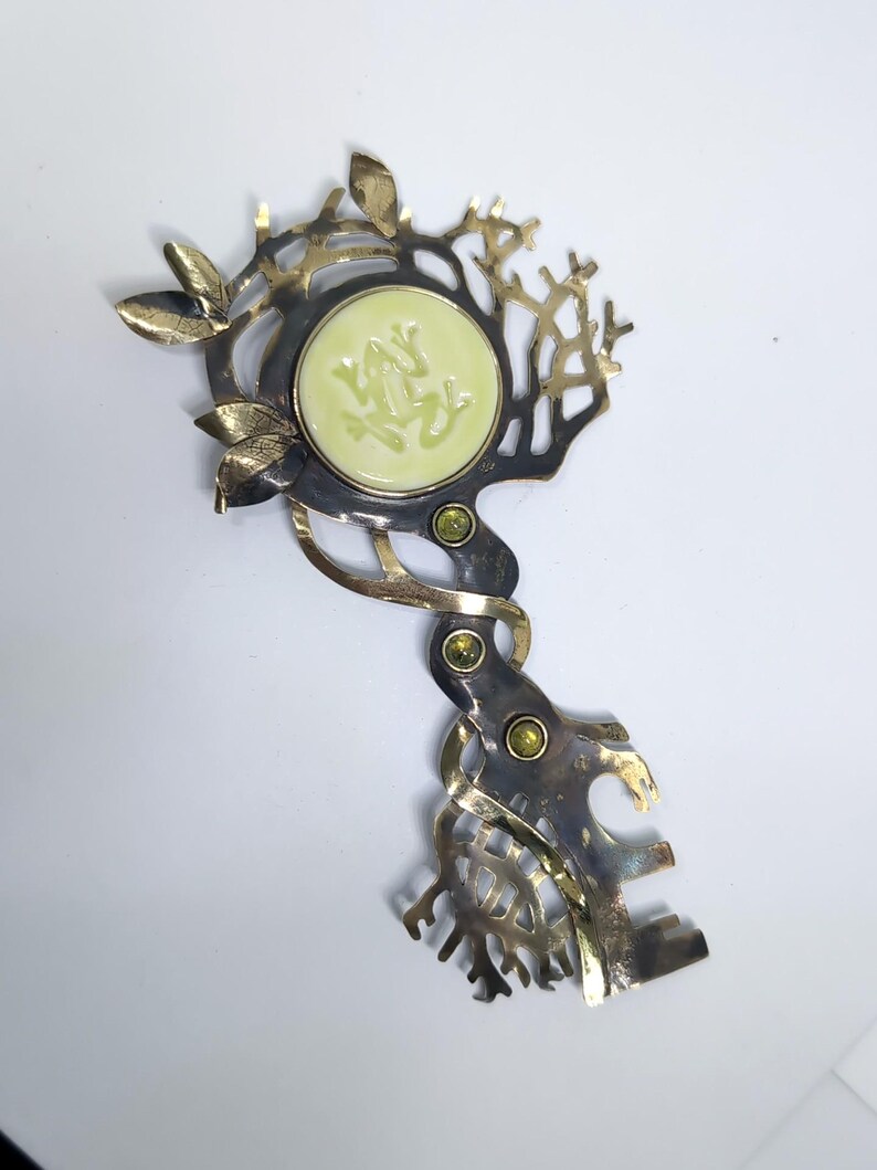 May include: A decorative metal key-shaped brooch featuring a central pale yellow circular inlay with a relief of a frog. The metalwork has a dark, oxidized finish with polished gold-tone leaf accents and three small, round, green inset gemstones.