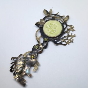 May include: A decorative metal brooch shaped like a key with a dark, oxidized finish. It features a central circular pale green inlay with a frog relief, three small green gemstones set along the stem, and intricate branch-like metalwork.