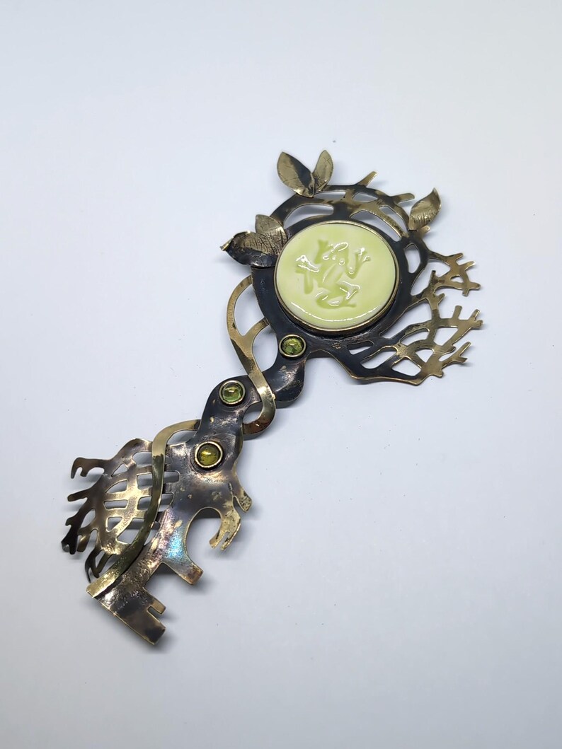 May include: A decorative metal brooch shaped like a key with a dark, oxidized finish. It features a central circular pale green inlay with a frog relief, three small green gemstones set along the stem, and intricate branch-like metalwork.