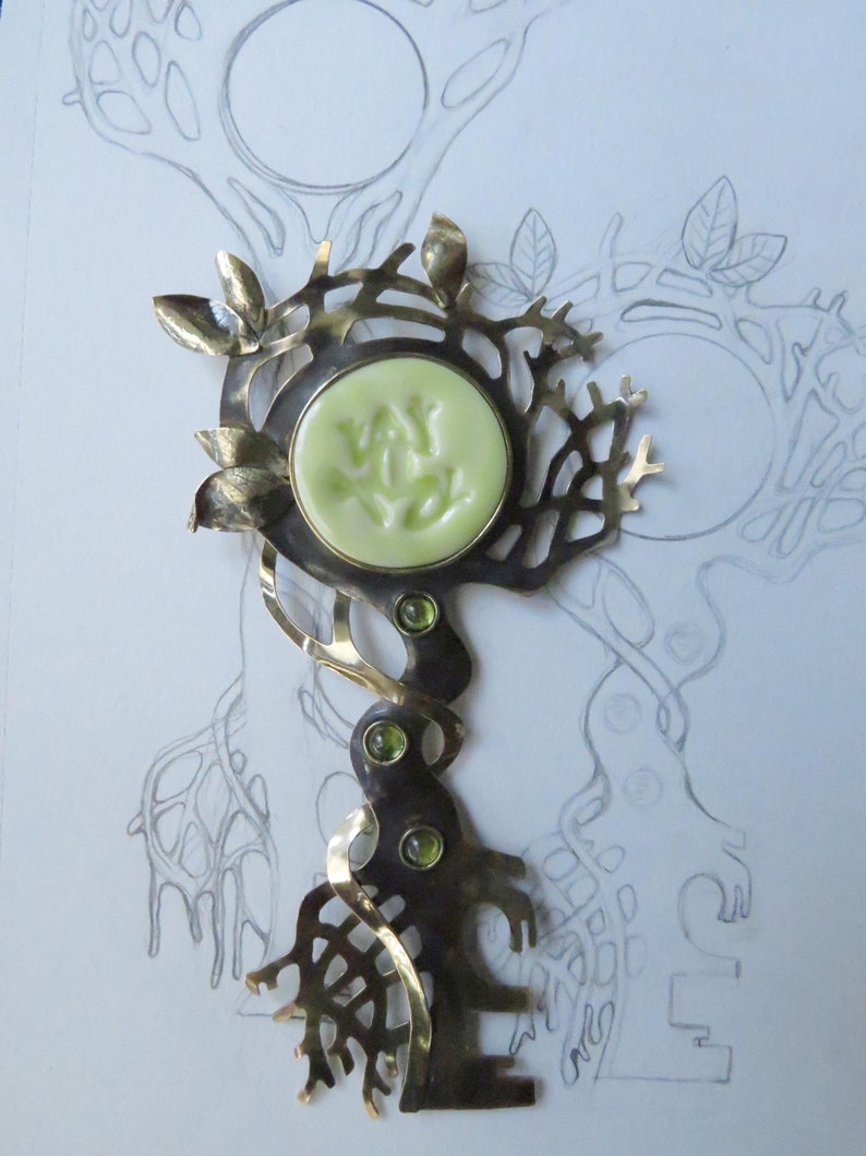 May include: A metal brooch shaped like a tree with a pale green circular center piece featuring embossed markings. The design includes metallic leaves, three small green inset stones along the trunk, and an intricate branch-like structure.