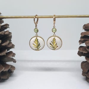 May include: A pair of gold-toned drop earrings featuring circular white pendants with a green fern leaf illustration. Each earring includes a small, round, light green gemstone accent above the pendant and a secure hinged hoop closure.