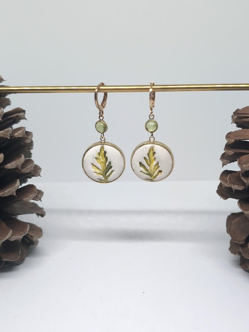 May include: A pair of gold-toned drop earrings featuring circular white pendants with a green fern leaf illustration. Each earring includes a small, round, light green gemstone accent above the pendant and a secure hinged hoop closure.